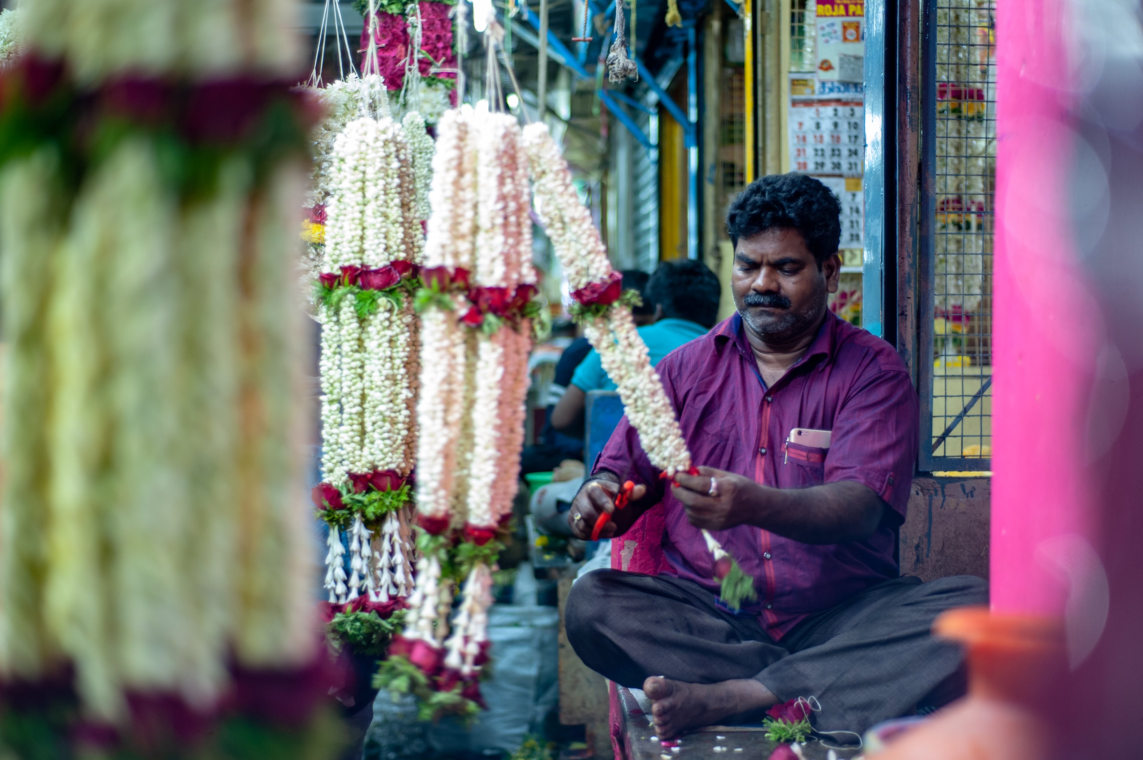 A flower vendor, sitting cross legged, works away on his orders for the day. 