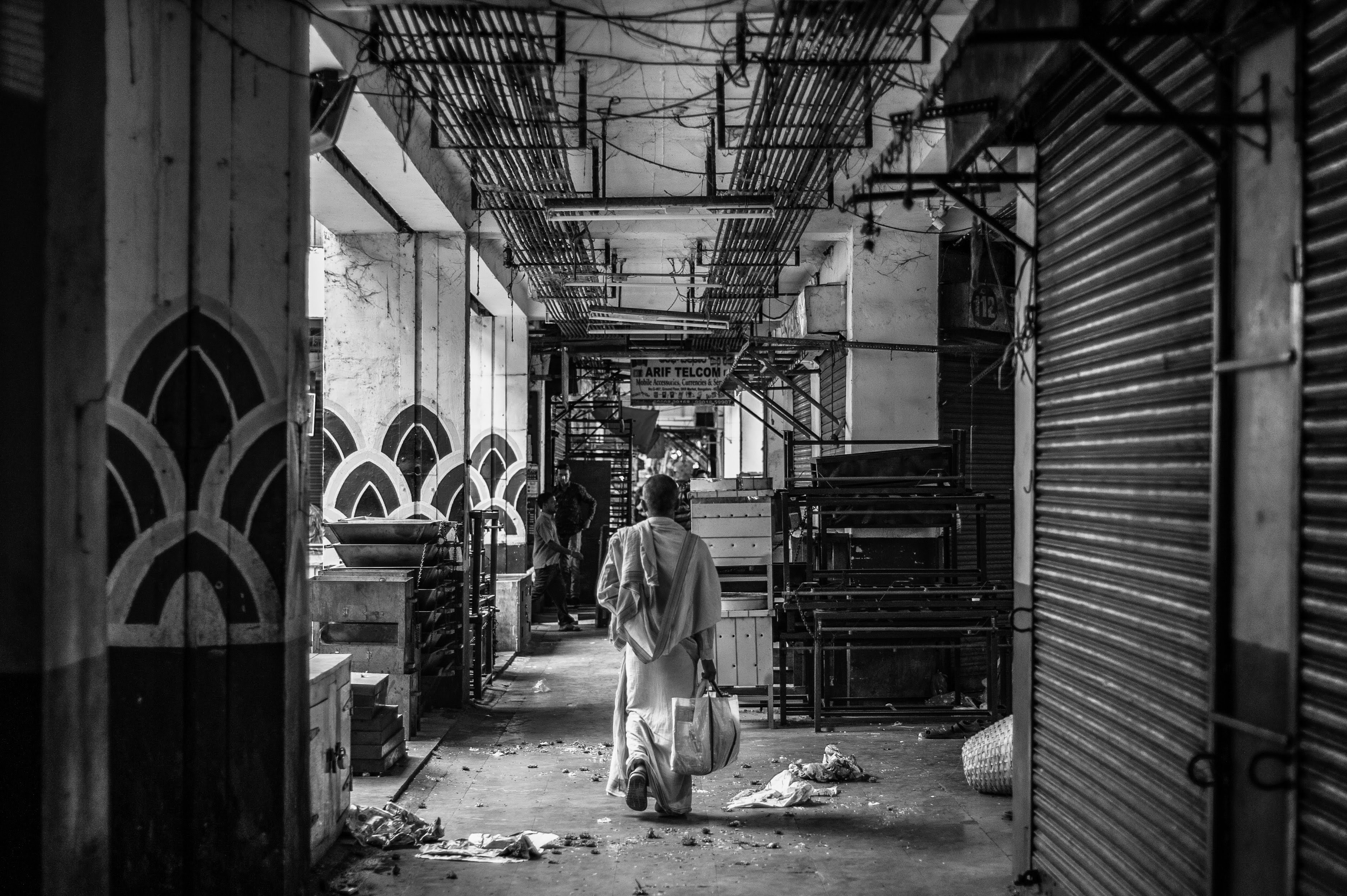 A black and white image of man carrying a plastic bag, walking through an empty KR Market in Bangalore. 